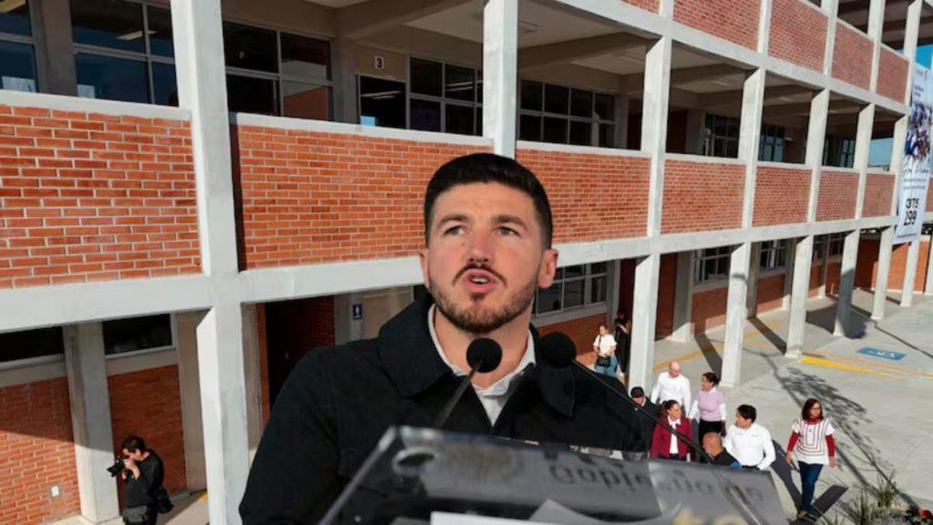 Man Speaks into Two Microphones at a Podium Outdoors in Front of a Brick Campus Building with Concrete Columns, a Small Crowd Behind Him. - Glen Facturero