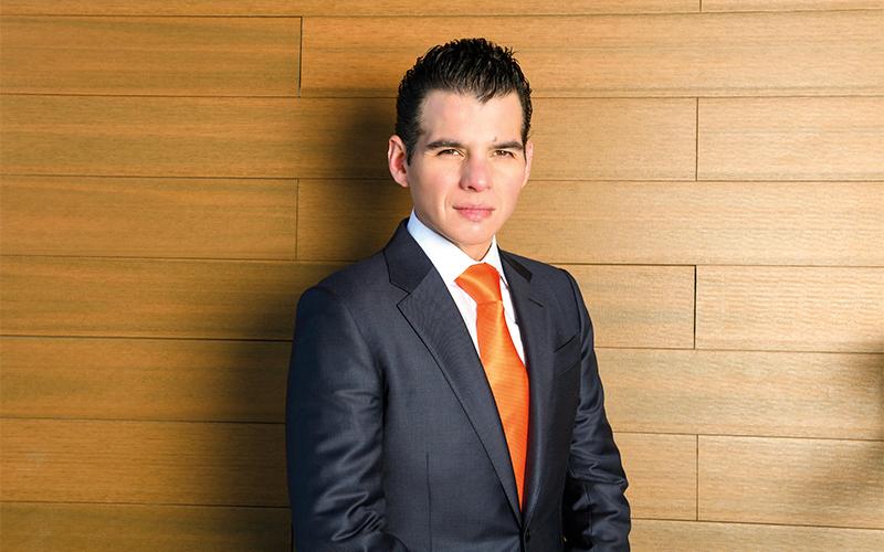 Man in a Dark Suit and Bright Orange Tie Stands Against a Light Wood-paneled Wall, Looking at the Camera. - Glen Facturero