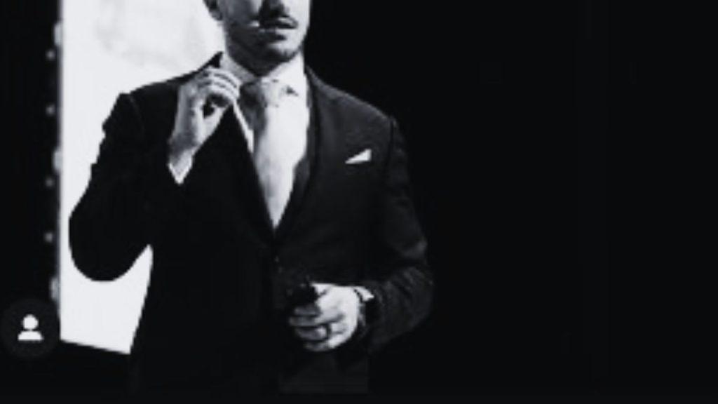 Man in a Dark Suit Adjusts His Tie, Holding a Small Device, Shown in Black and White. - Glen Facturero