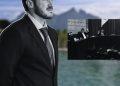 Man in a Dark Suit and Tie Stands in the Foreground Outdoors by a Blurred Lake, with a Black-and-white Inset of a Formal Meeting Panel Raising Hands. - Glen Facturero
