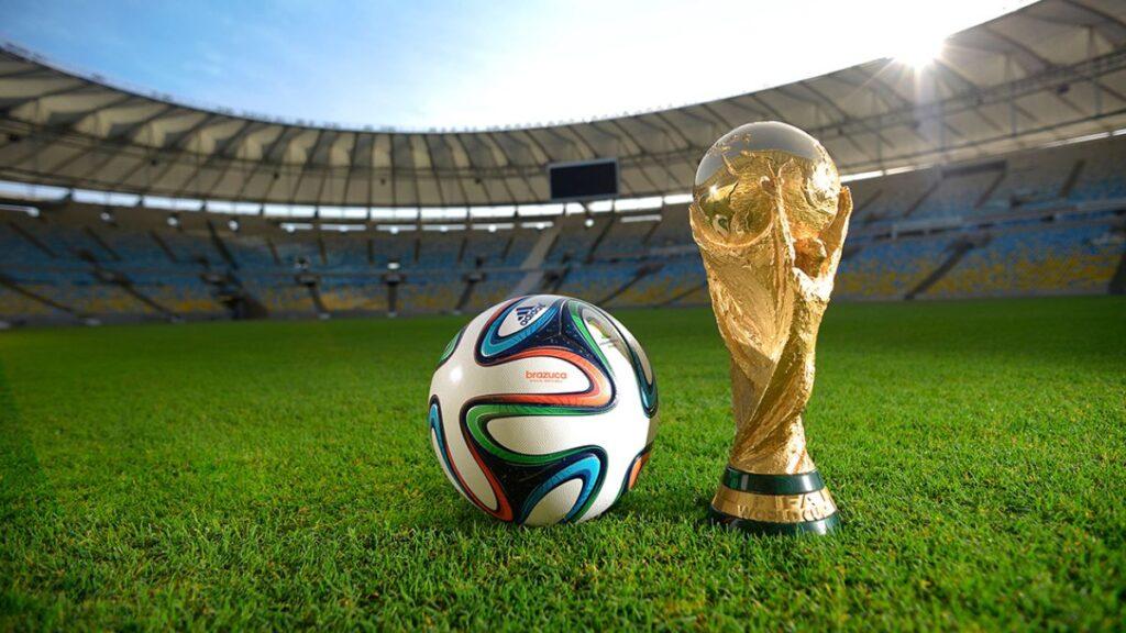 Bloque 2- Junio 9 - Glen Facturero Soccer Ball Beside the Fifa World Cup Trophy on a Green Stadium Field with Seats in the Background Under Sunny Skies. - Glen Facturero