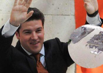 Smiling man in a dark suit and orange tie waves hello, with a circular inset showing a muddy construction site nearby.