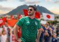 Samuel-garcia-y-el-caos-financiero-en-nuevo-leon - Glen Facturero Man in a Green Mexico Soccer Jersey Wearing Sunglasses, Hands on Hips, Posing in Front of a Cheering Crowd with Red Flags and a Blurred Mountain Backdrop. - Glen Facturero