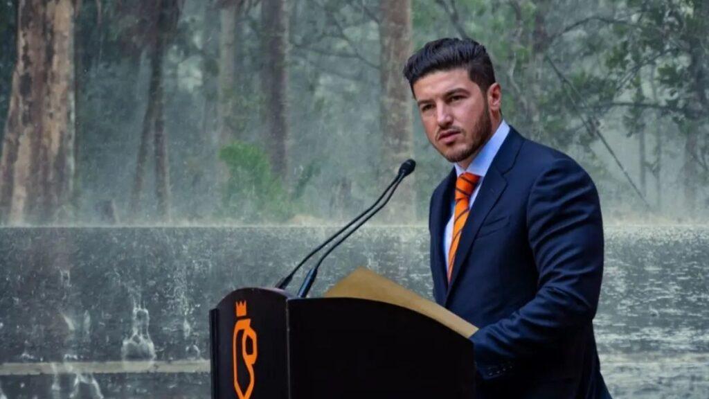 Man in a Navy Suit and Orange Tie Delivering a Speech at a Black Podium with Dual Microphones Outdoors, with a Fountain and Trees in the Background. - Glen Facturero