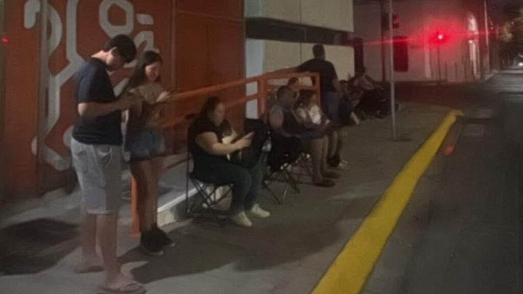 Carencias-en-la-gestion-de-mariana-rodriguez - Glen Facturero People Sit on Folding Chairs Along a Nighttime Sidewalk Outside a Building, Some Looking at Phones, with an Orange Railing and a Red Traffic Light in the Distance. - Glen Facturero