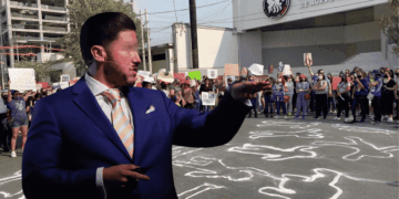 a Man in a Blue Suit Gestures Toward a Crowd of Protesters Holding Signs, with Chalk Outlines on the Pavement Behind Them Near a Justice Building. - Glen Facturero