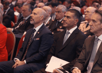 Audience at a Formal Event: Three Men in Suits Seated in the Foreground, with a Woman in Red on the Left and Others in the Background. - Glen Facturero