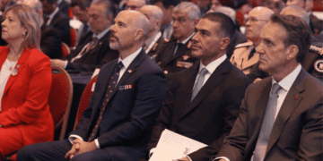 Audience at a Formal Event: Three Men in Suits Seated in the Foreground, with a Woman in Red on the Left and Others in the Background. - Glen Facturero