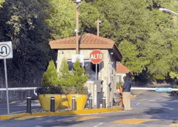 Small security booth at a gated entrance with a red 'ALTO' stop sign, barrier gate, and yellow planters nearby.