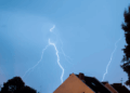 Lightning Bolts Strike Across a Blue Sky over a Neighborhood with Pitched Roofs and Trees on Both Sides. - Glen Facturero