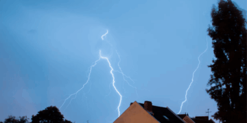 Lightning Bolts Strike Across a Blue Sky over a Neighborhood with Pitched Roofs and Trees on Both Sides. - Glen Facturero
